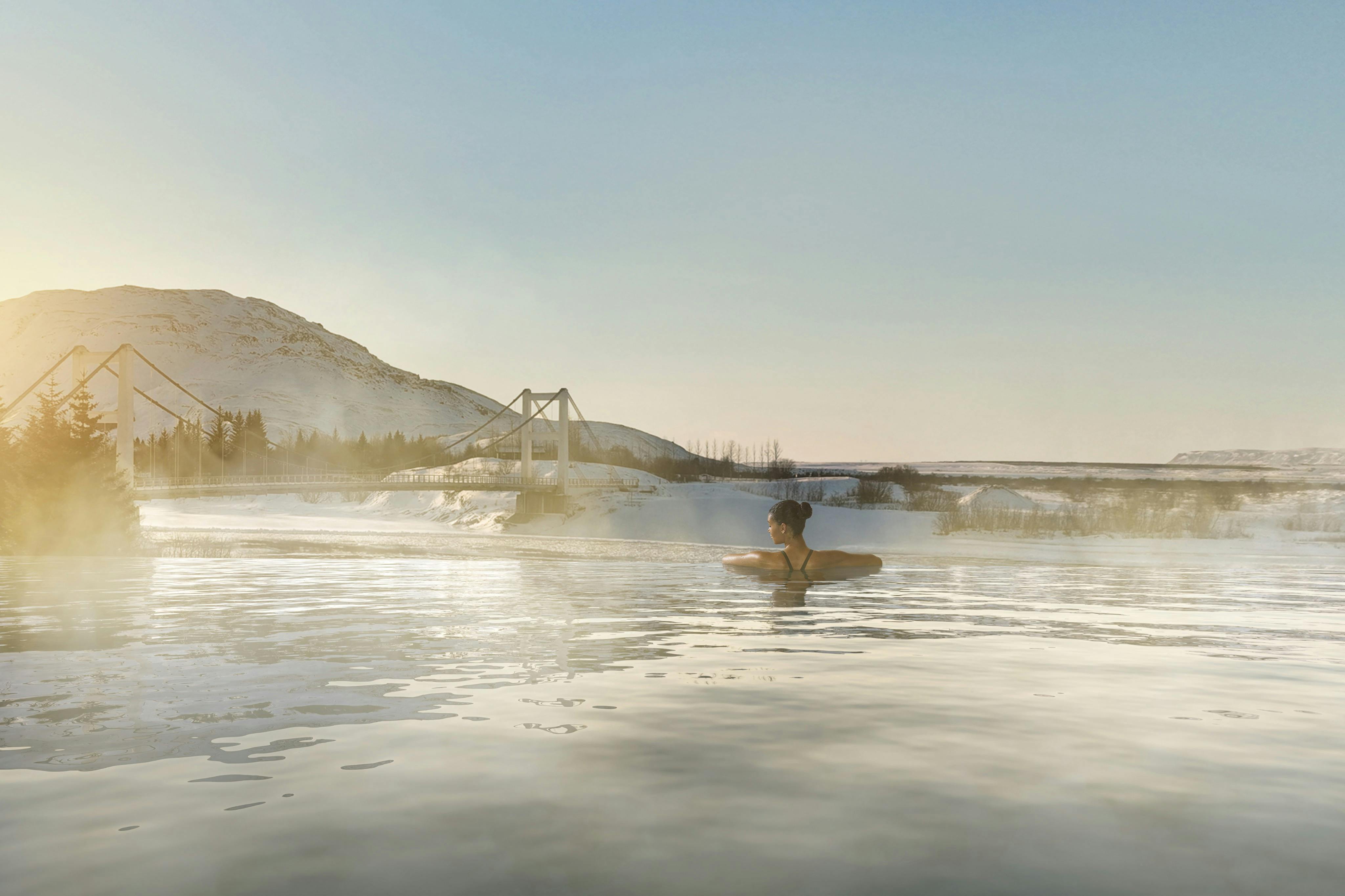Person relaxes in a calm hot spring with snow-covered landscape, hills, and a bridge in the background under a clear sky.