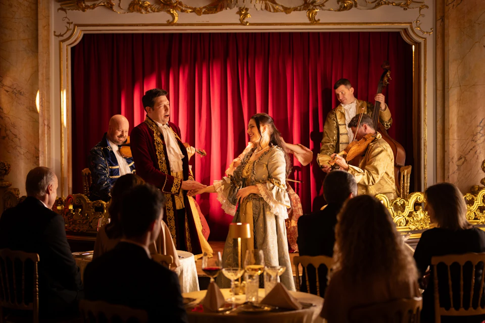 A man and woman in period costumes perform in front of a red curtain, while seated diners watch in an ornate setting.
