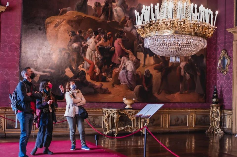 Two masked women observe a large historical painting in an ornate museum room with a chandelier and a red rope barrier.