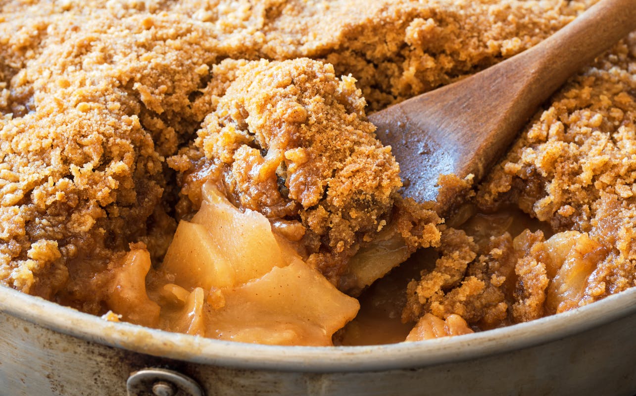 Close-up of a serving of apple crumble being scooped with a wooden spoon, revealing baked apples and crumbly topping.
