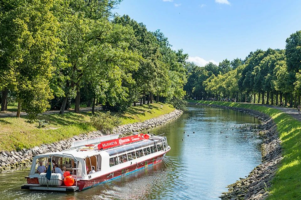 A red and white tour boat navigates a tree-lined canal under a clear blue sky. Ducks are visible in the water ahead.