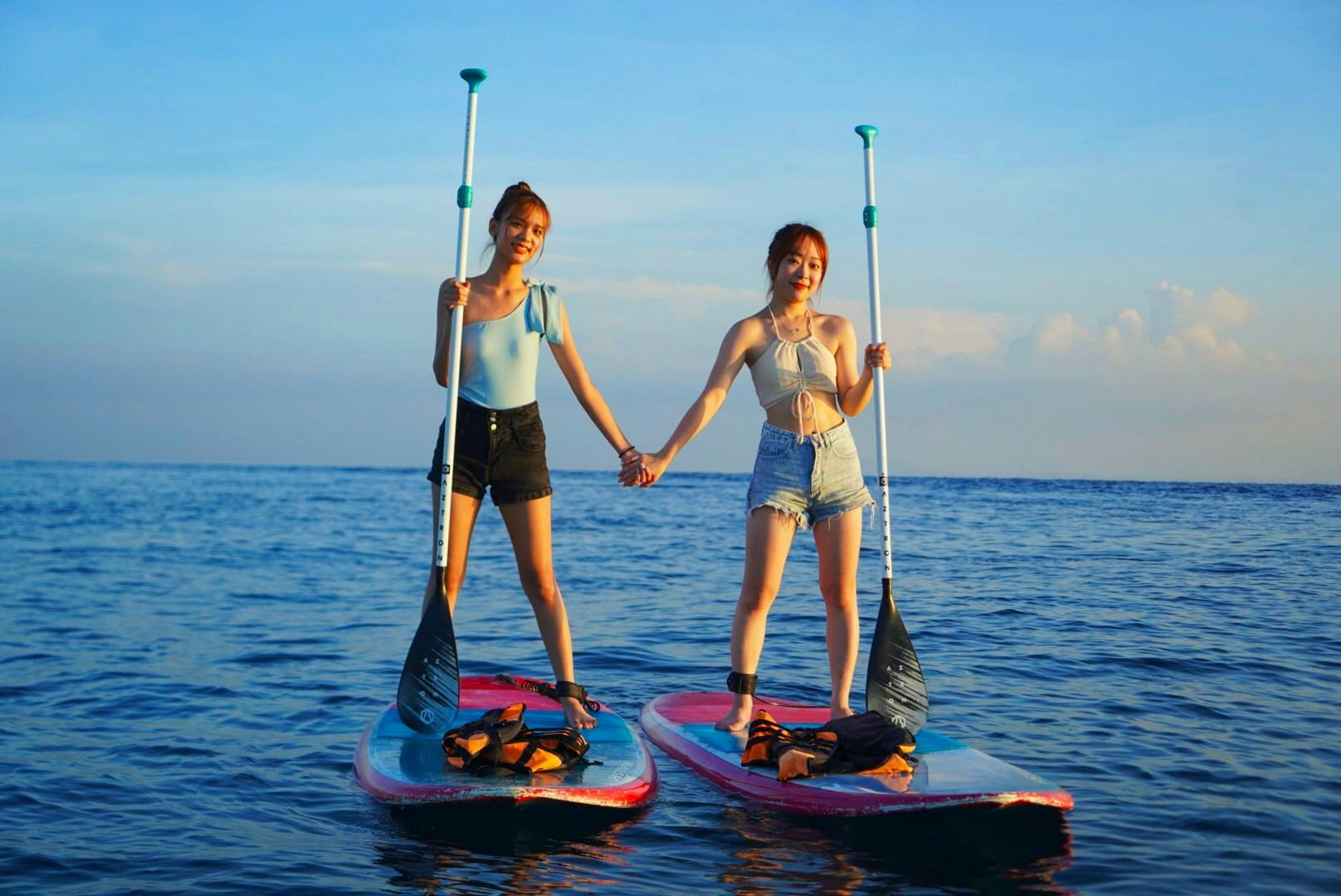 Two people standing on paddleboards in a calm sea, holding hands and smiling, under a clear blue sky during sunset.