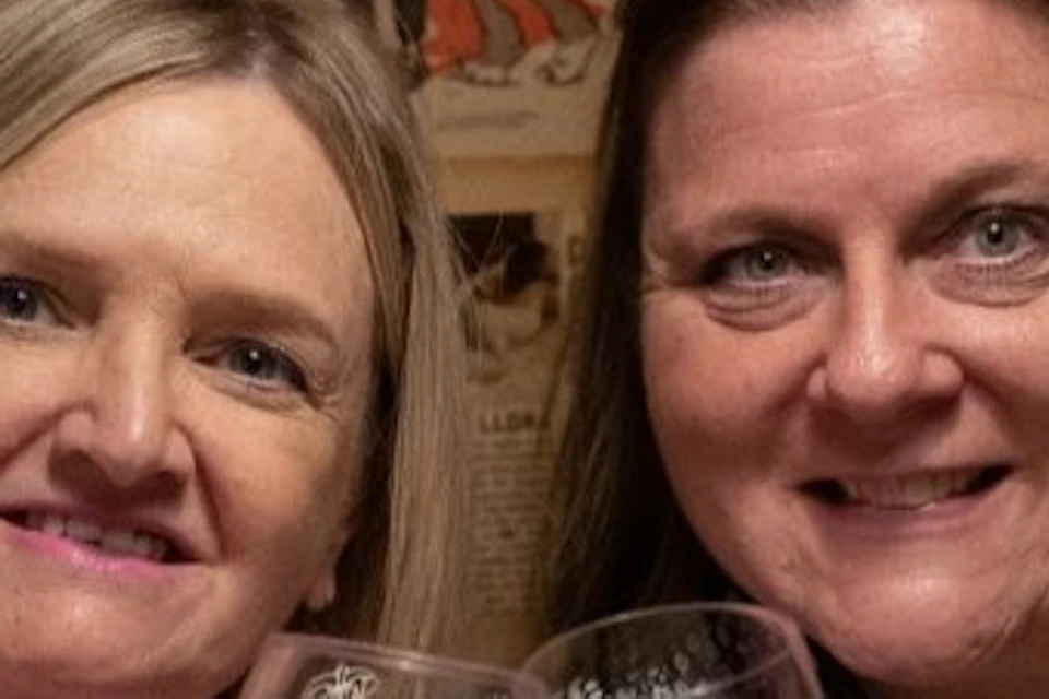Two women sitting at a table holding glasses of red wine, with a wall decorated with vintage posters behind them.