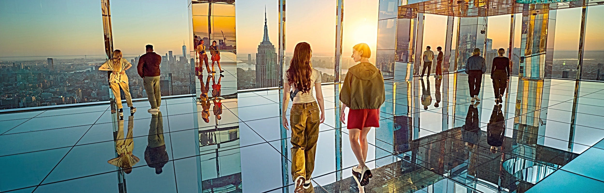 People stand and walk on a mirrored observation deck with a view of a city skyline at sunset.
