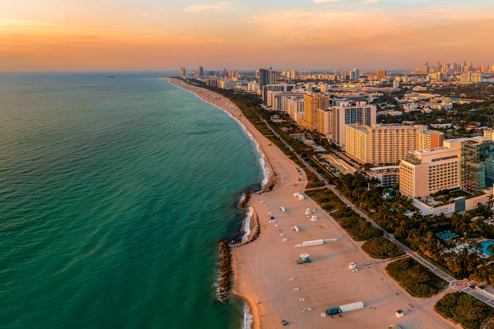 Aerial view of a coastline with beige sandy beach, turquoise water, and numerous high-rise buildings under a partly cloudy sky.