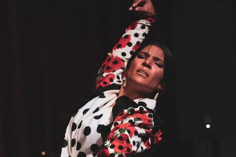 Person in a polka-dot and floral-patterned outfit dancing, arm raised, with a focused expression in low lighting.