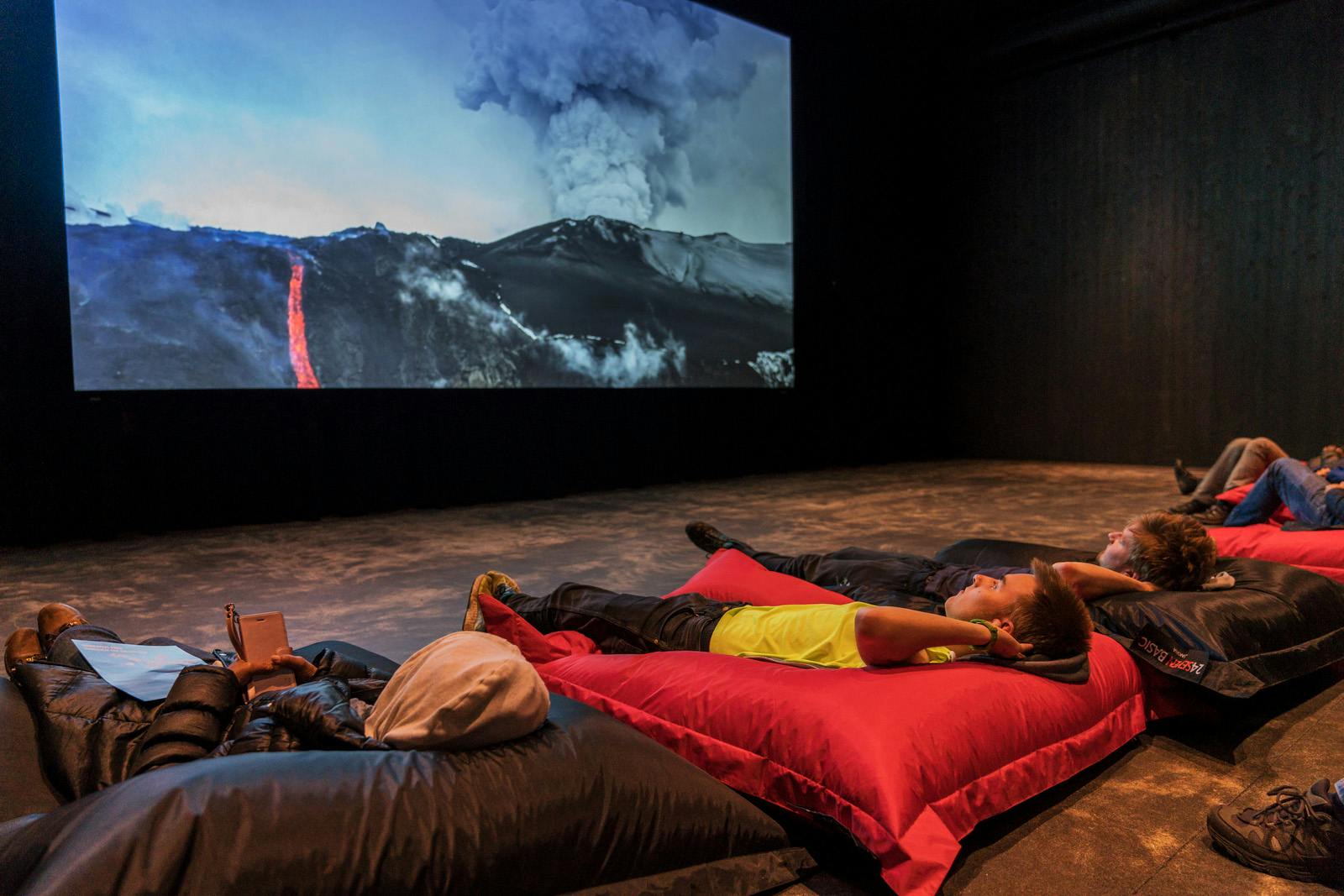 어두운 방의 커다란 빨간 쿠션에서 휴식을 취하는 사람들은 대형 화면에서 화산 폭발을 목격합니다.
