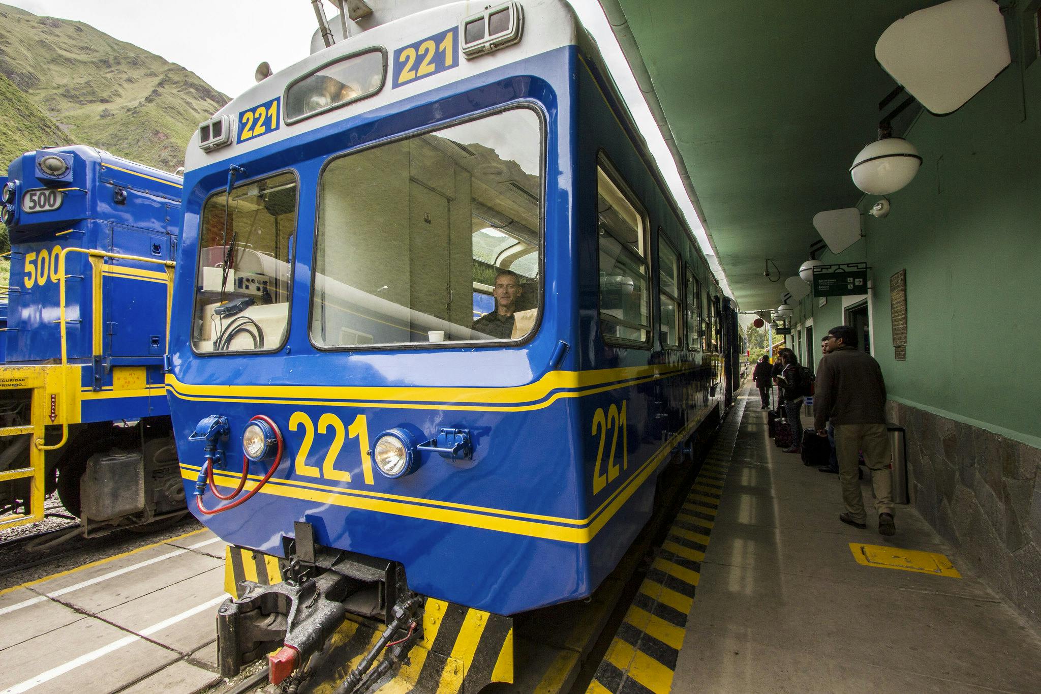 Ένα φωτεινό μπλε τρένο με κίτρινα σημάδια με την ένδειξη "221" σταθμεύει σε μια πλατφόρμα. Ένας αγωγός είναι ορατός από το παράθυρο.