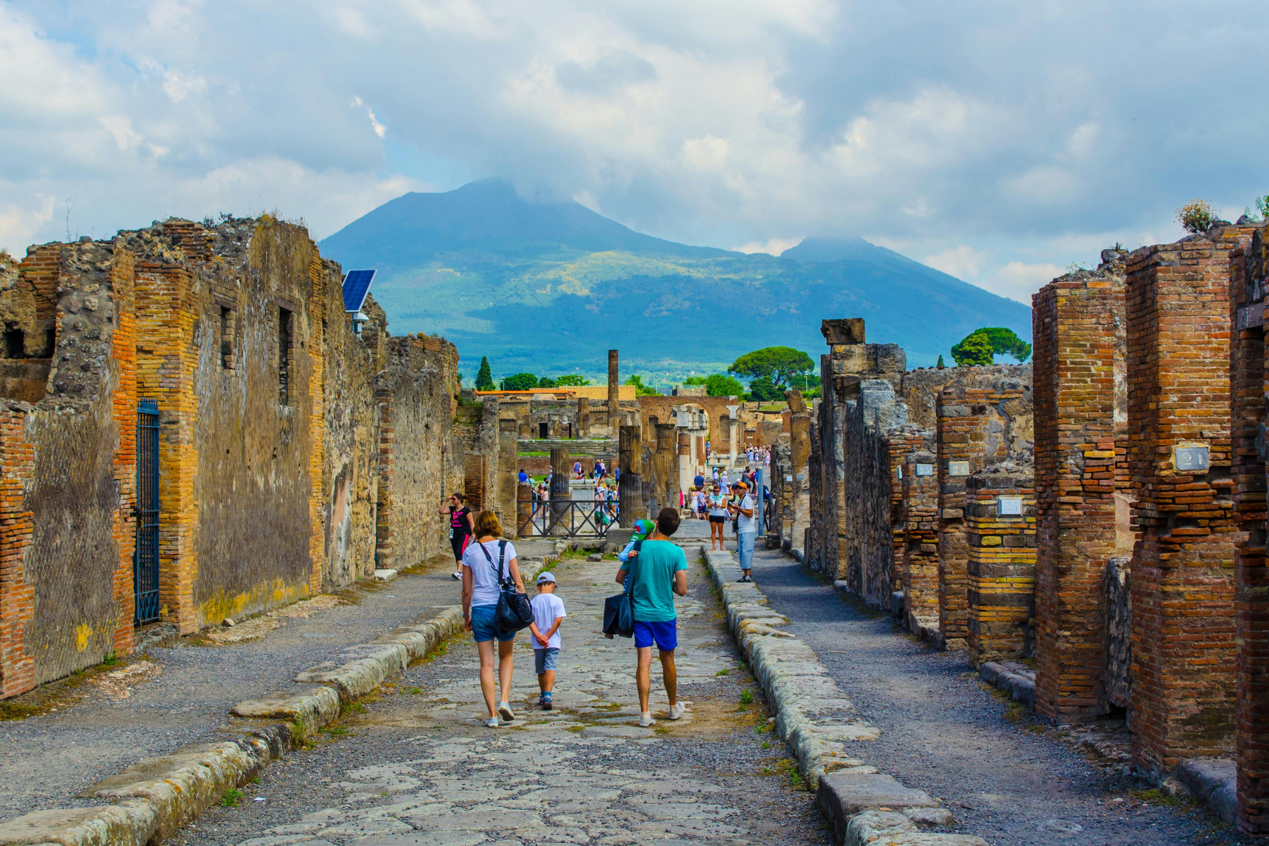 Tourists walk through the ancient ruins of Pompeii, with Mount Vesuvius visible in the background under a partly cloudy sky.