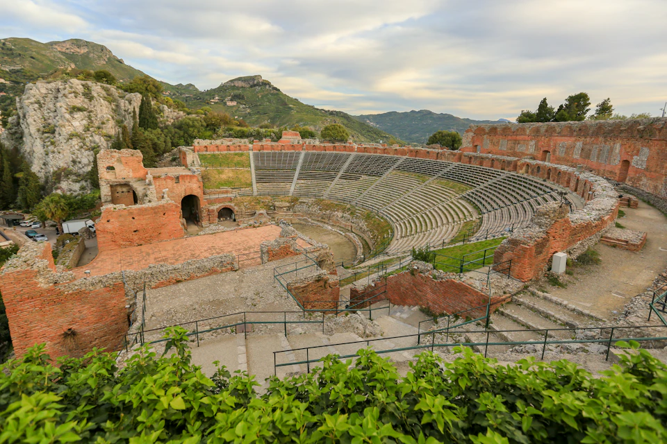 Ancient amphitheater with tiered stone seating and surrounding ruins, set against a backdrop of green hills and a cloudy sky.