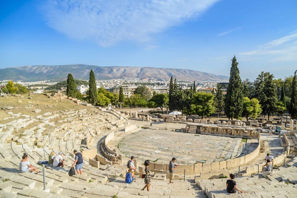 A historical amphitheater with stone seating and a circular stage, surrounded by trees and hills under a clear sky.