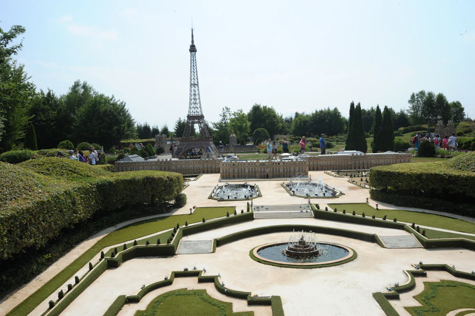 A scale model Eiffel Tower in a manicured garden setting with pathways, fountains, and miniature buildings, surrounded by greenery and tourists.