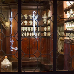 Ett träskåp fyllt med flera hyllor av porslinsburkar och vaser, sedda genom ett blyglasfönster.