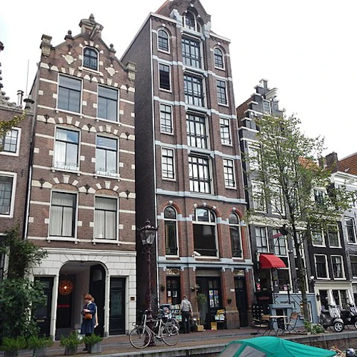 Rad av traditionella holländska kanalhus med utsmyckade fasader i Amsterdam. Cyklar och fotgängare syns framför.