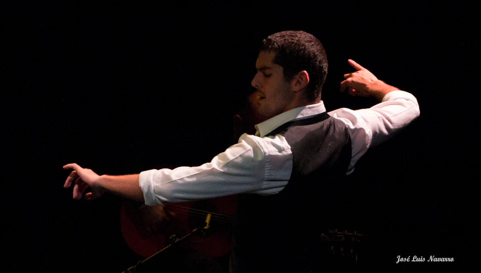 Man in white shirt and dark vest, mid-dance, against a dark background with a partially visible guitar and musician.