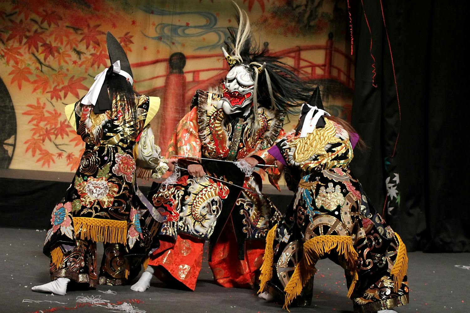 Performers in ornate costumes and masks during a traditional Japanese dance on stage with a painted backdrop.