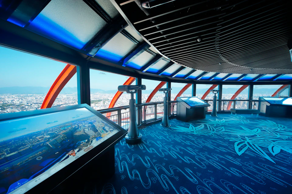 Observation deck with telescopes and interactive touch screens, offering panoramic city views through large windows with blue accents.