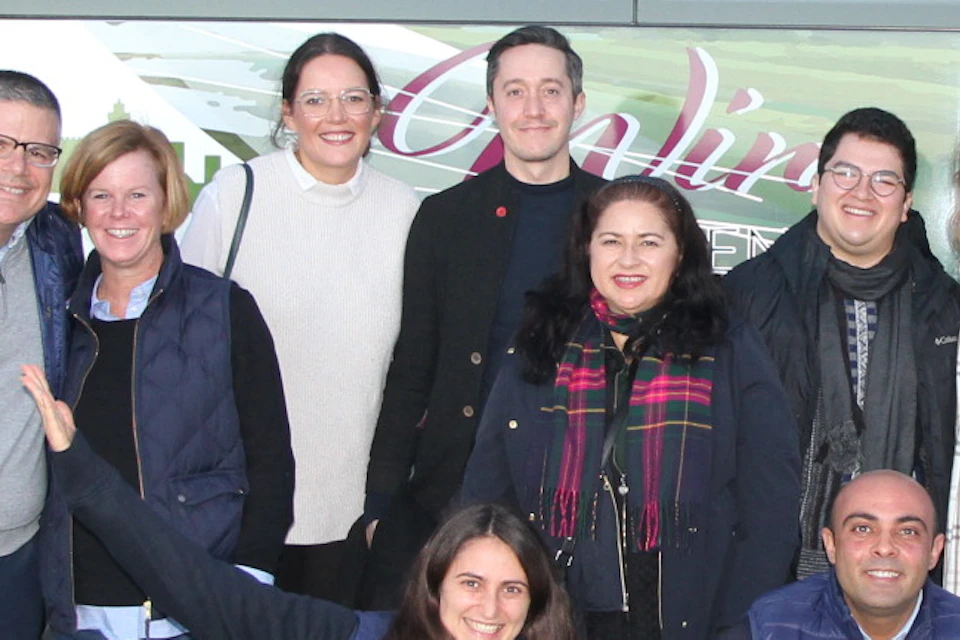 Group of 14 people posing in front of a parked tour bus on a clear day. Most are smiling and wearing casual clothes.