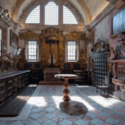 Utsmyckat rum med välvda fönster, detaljerade träarbeten, antika möbler, förgyllda dekorationer och ett krucifix på ett altare.