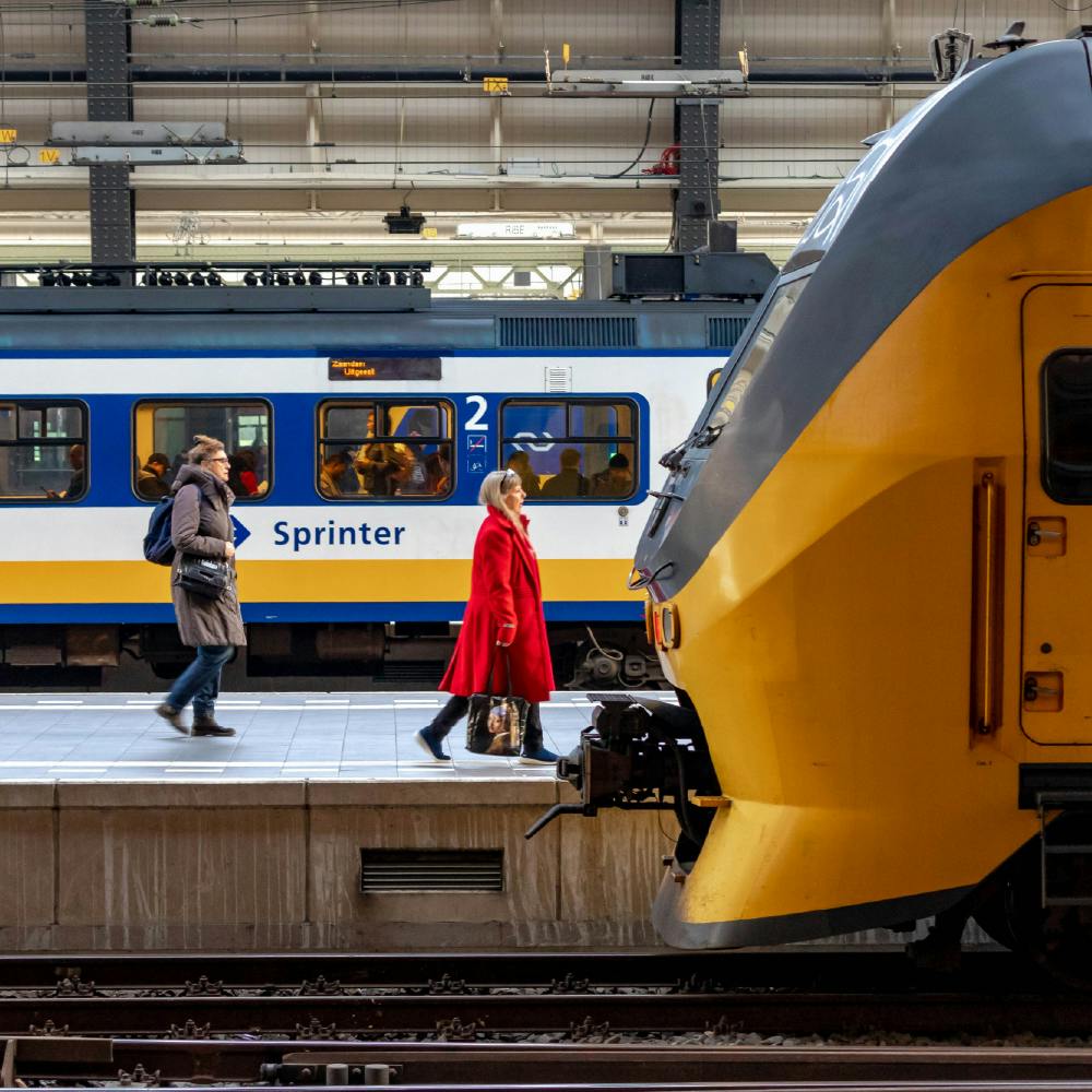 Due persone camminano su una piattaforma davanti a un treno giallo. Un altro treno con la scritta "Sprinter" è sullo sfondo.