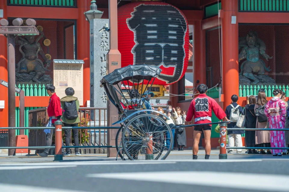 A traditional rickshaw in front of a large red lantern and temple entrance, with people walking around.