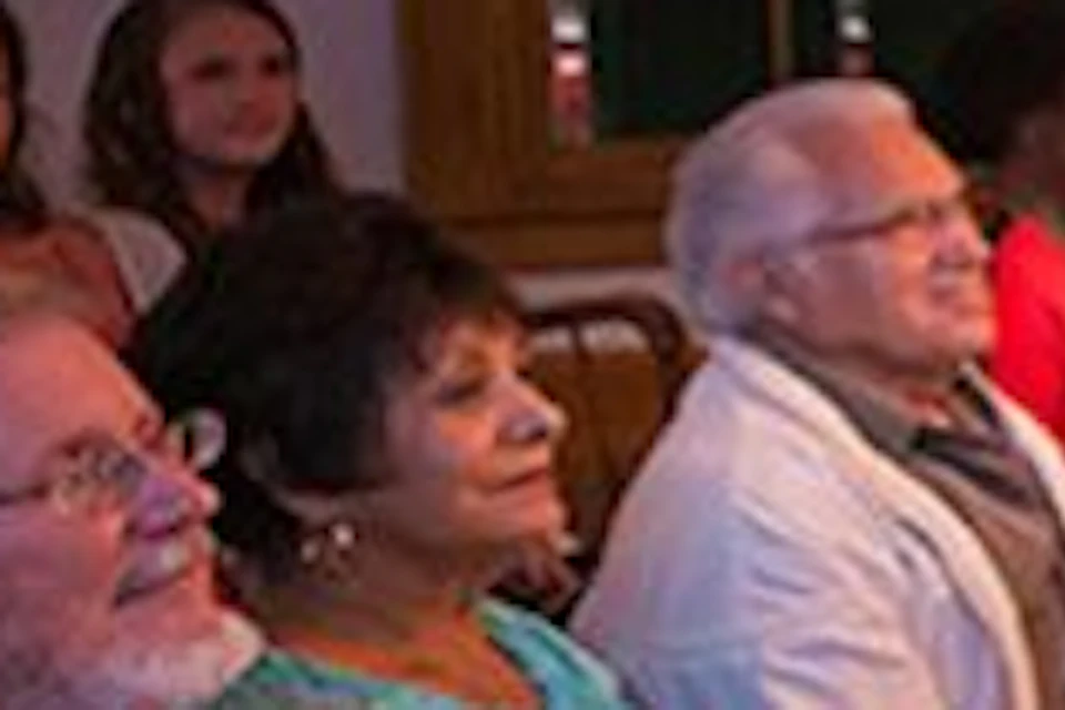 People seated in rows in a dimly lit room, listening to a woman in period attire speaking, with others observing from behind a railing.