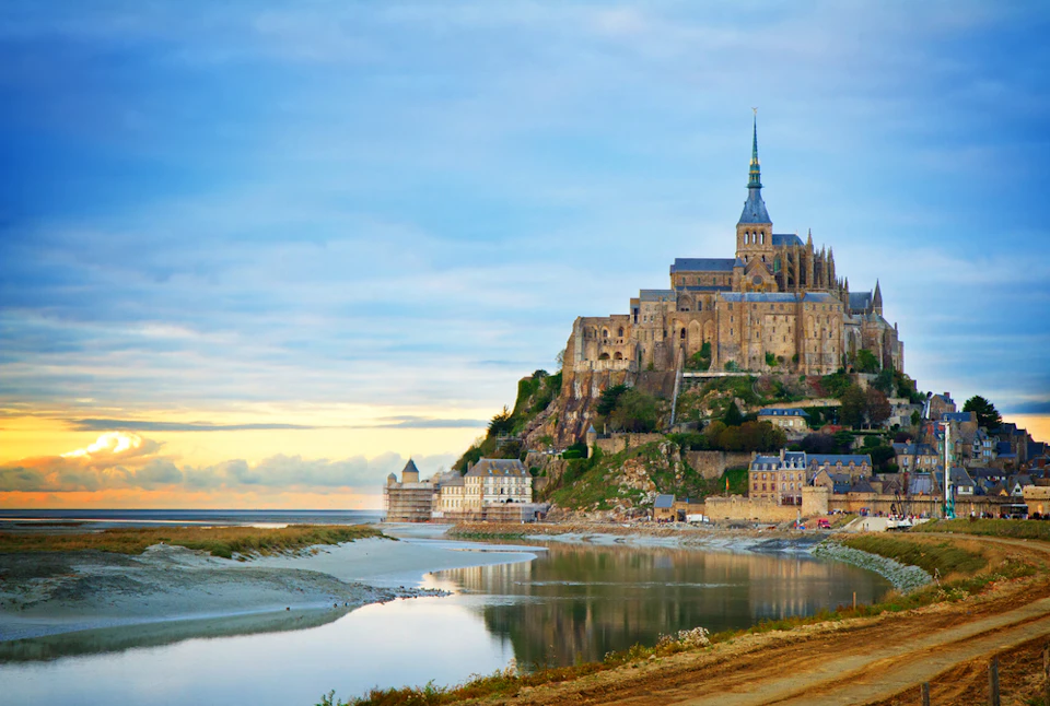 Medieval abbey and buildings on a rocky island surrounded by water, with a bridge connecting to the mainland at sunset.