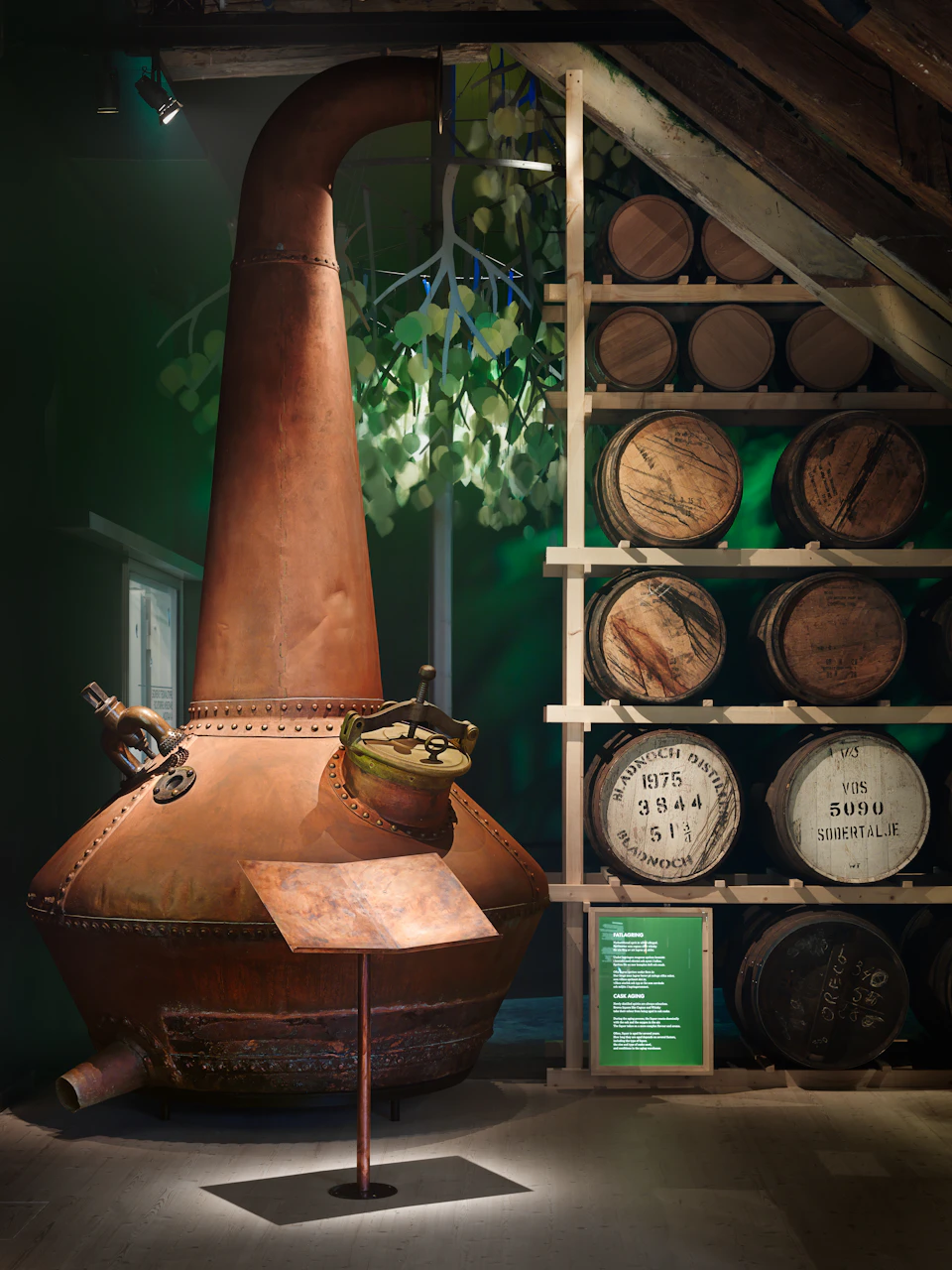 A copper distillation kettle sits next to a rack of aging wooden barrels in a cellar with foliage visible in the background.