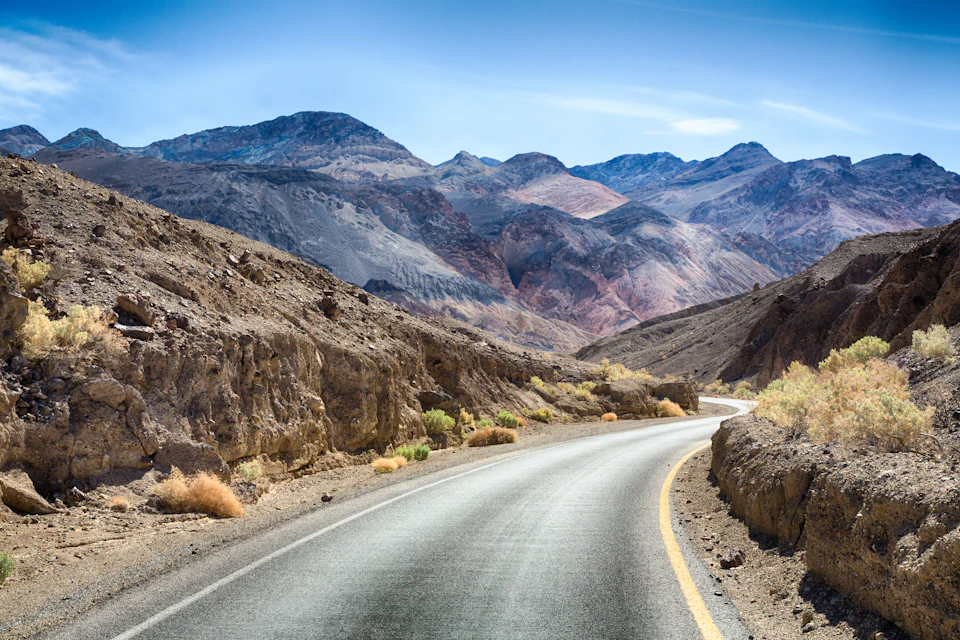 A winding road through a rocky desert landscape with mountains in the background under a clear blue sky.