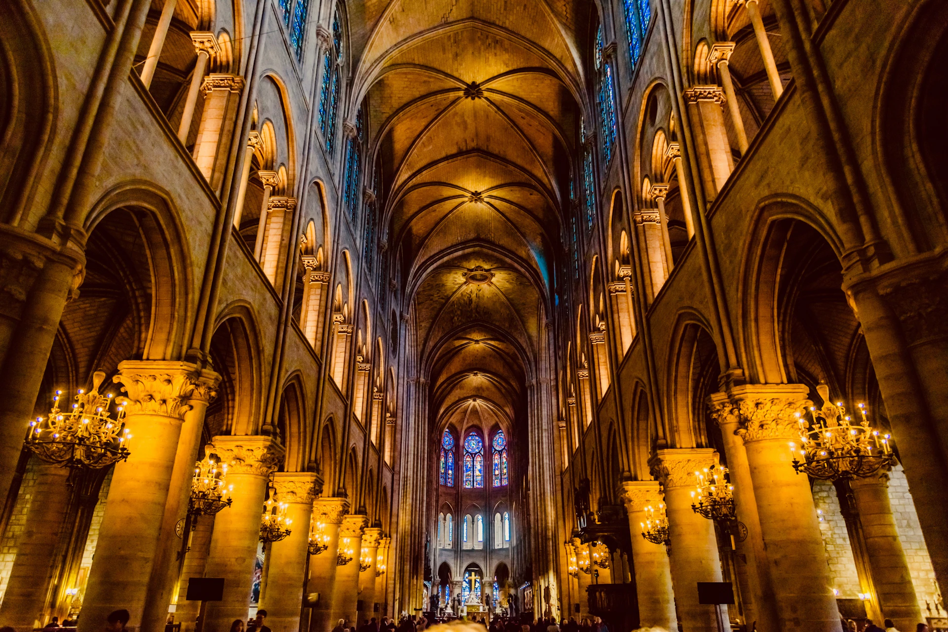 Interior of a grand cathedral with high vaulted ceilings, ornate columns, glowing chandeliers, and stained glass windows in the background.