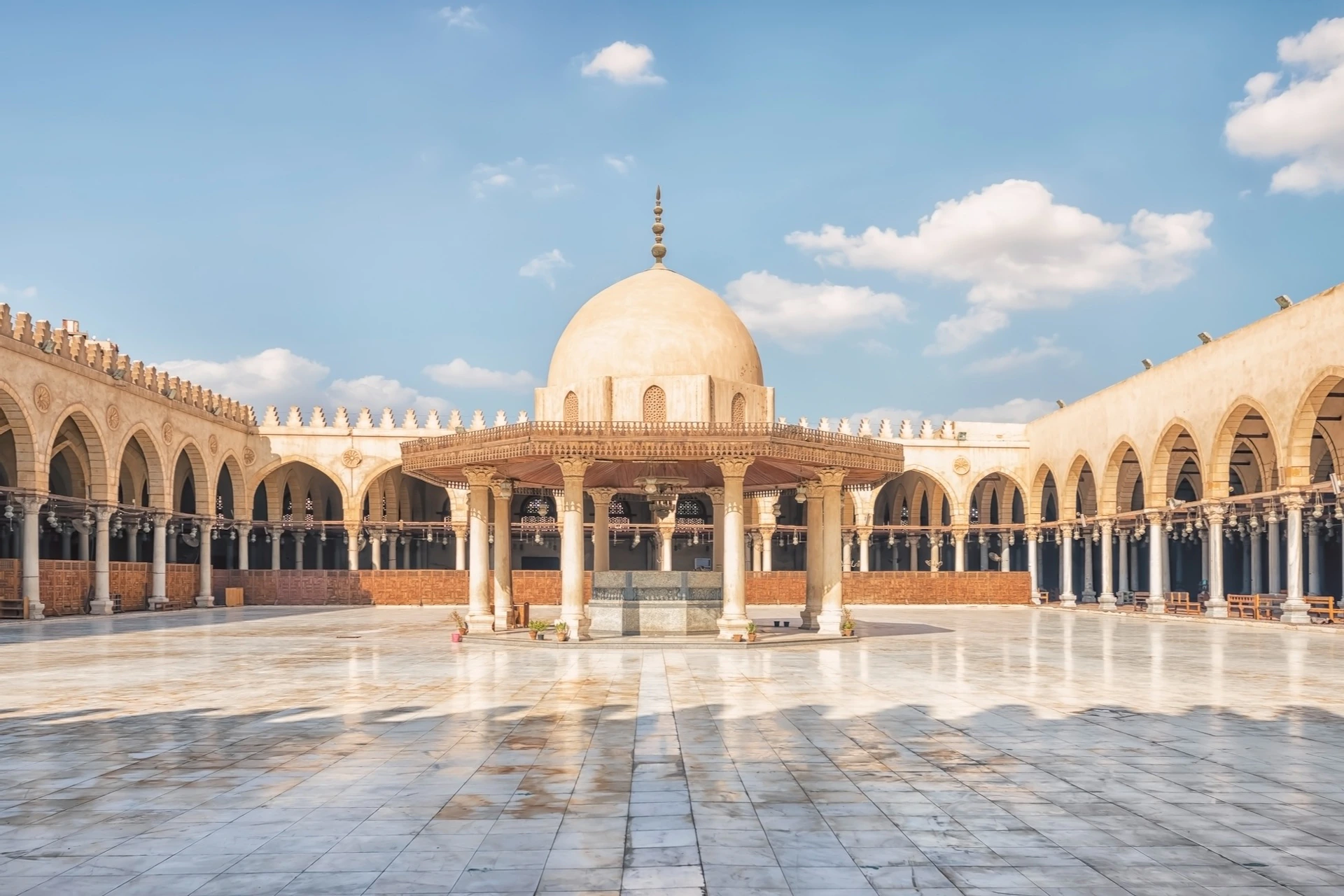 A mosque courtyard with a central dome structure, surrounded by arches and columns, under a blue sky with a few clouds.