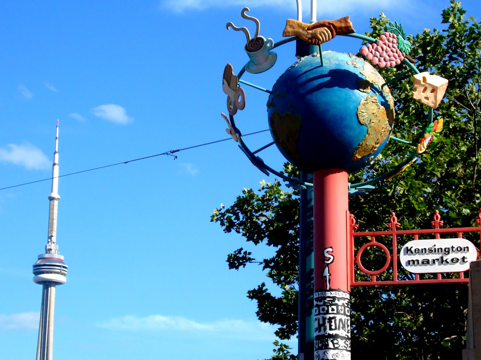 A signpost with a globe and various sculptures, including a coffee cup and pretzel, against a blue sky and tree background.