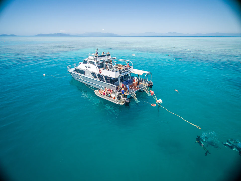 A boat anchored in clear blue water with people snorkeling nearby and a small boat attached at the rear.