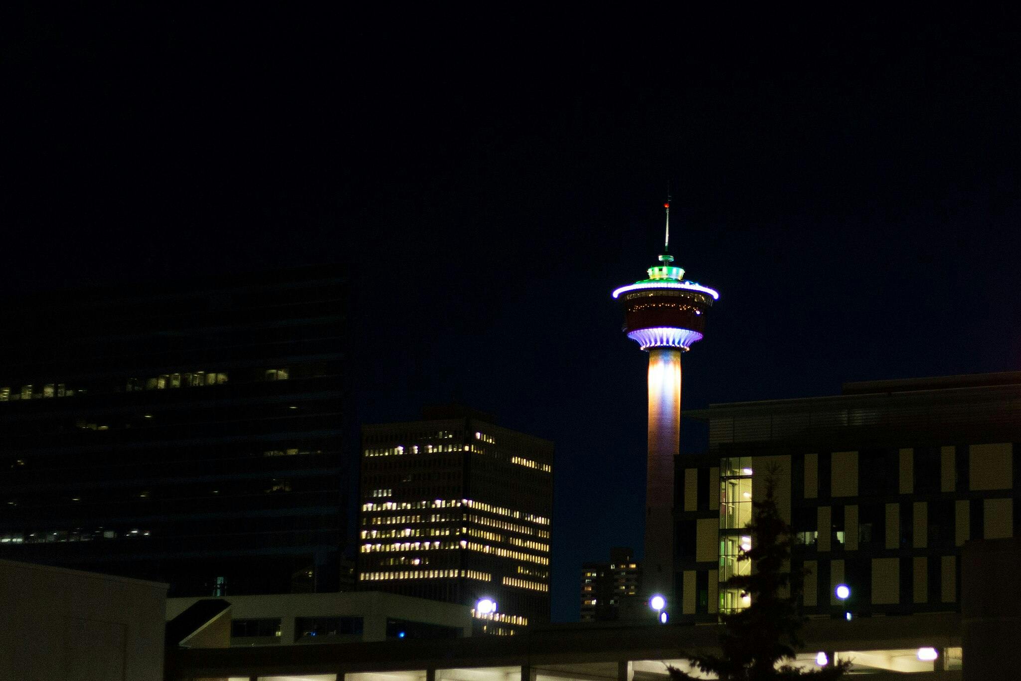 A brightly lit tower stands among buildings at night, with colorful lights illuminating the structure against the dark sky.