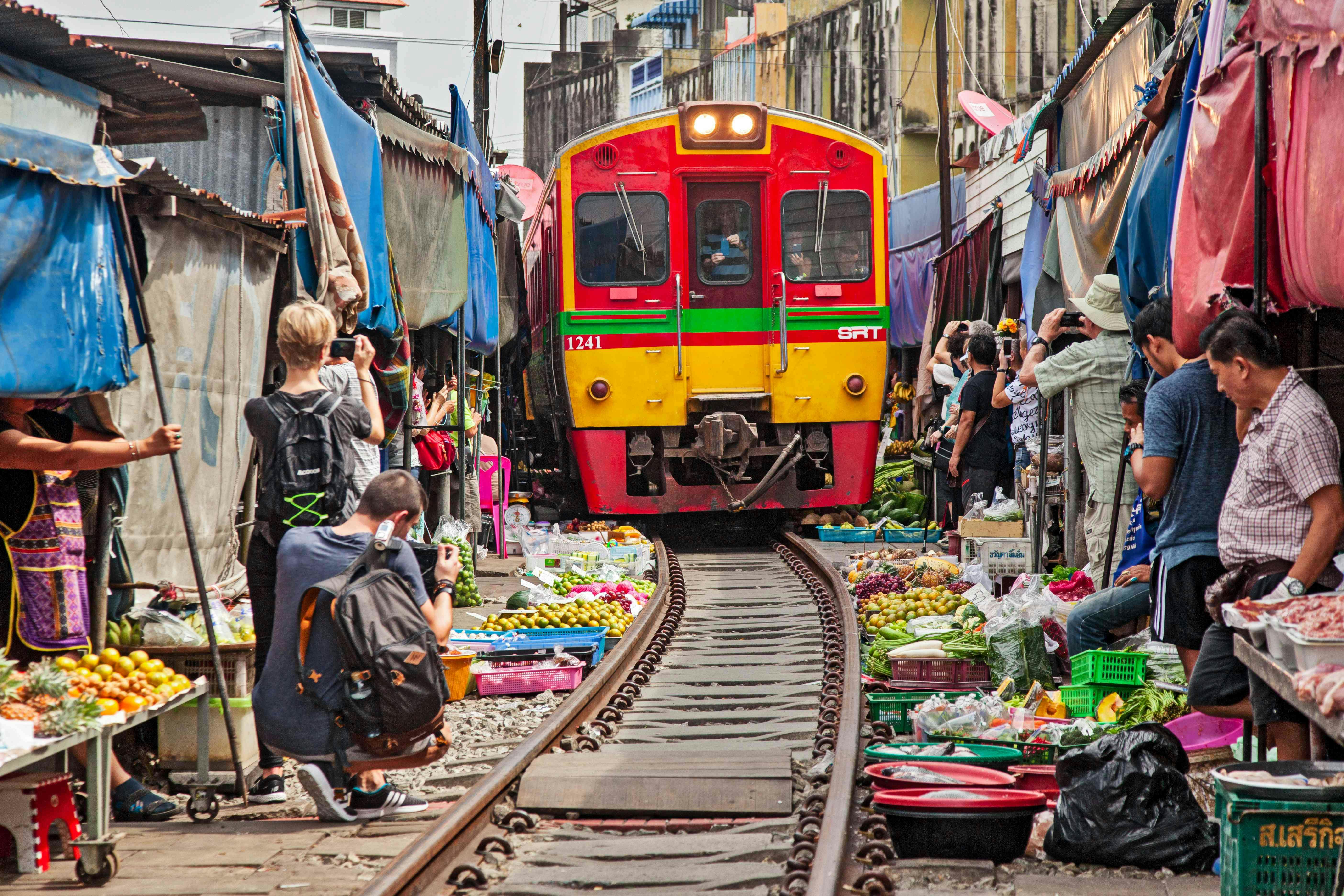 A colorful train passing through a crowded market street, with vendors and visitors close to the tracks taking photos.