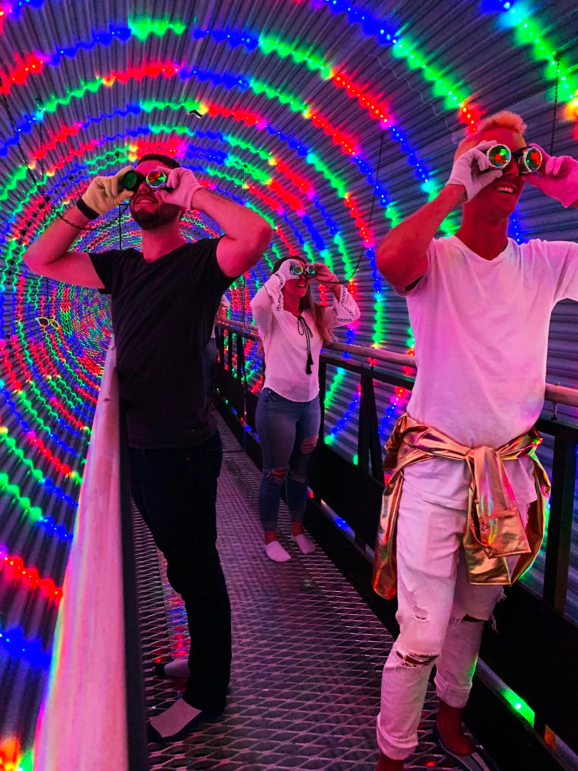 People stand on a walkway inside a tunnel illuminated by colorful LED patterns, wearing goggles and observing the lights.