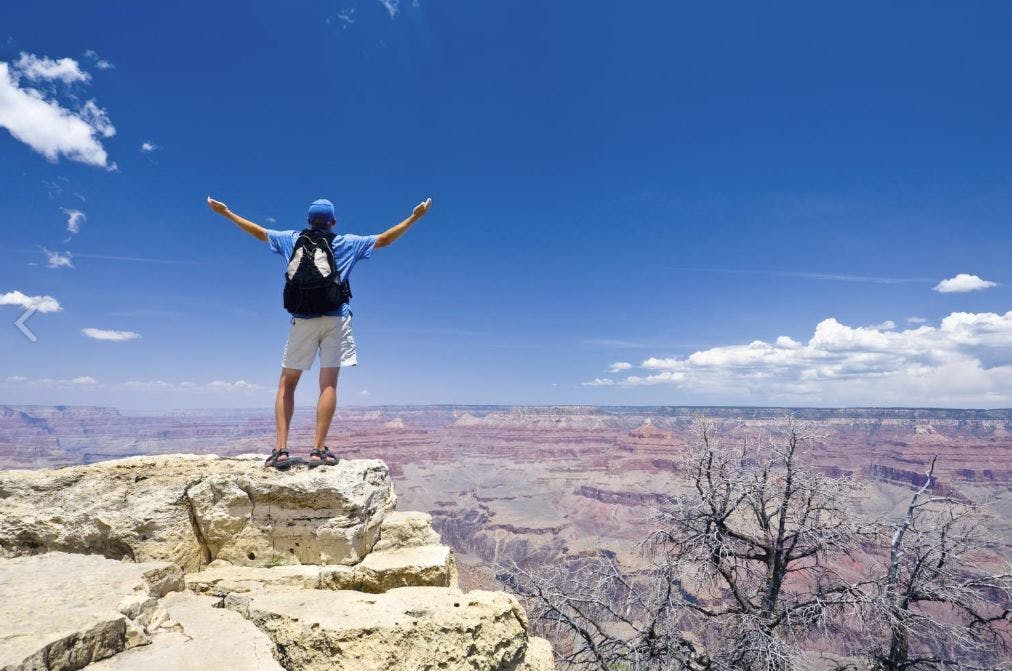 Person with a backpack stands on a rocky ledge with arms outstretched, overlooking a vast canyon under a clear blue sky.