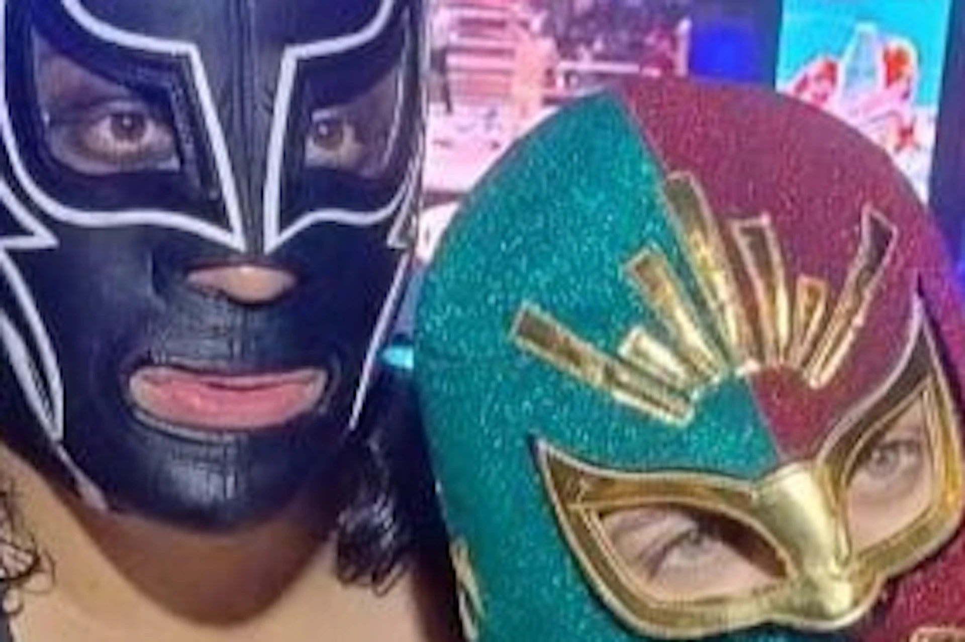 Two people in colorful wrestling masks pose in an arena with bright lights and a large screen in the background.