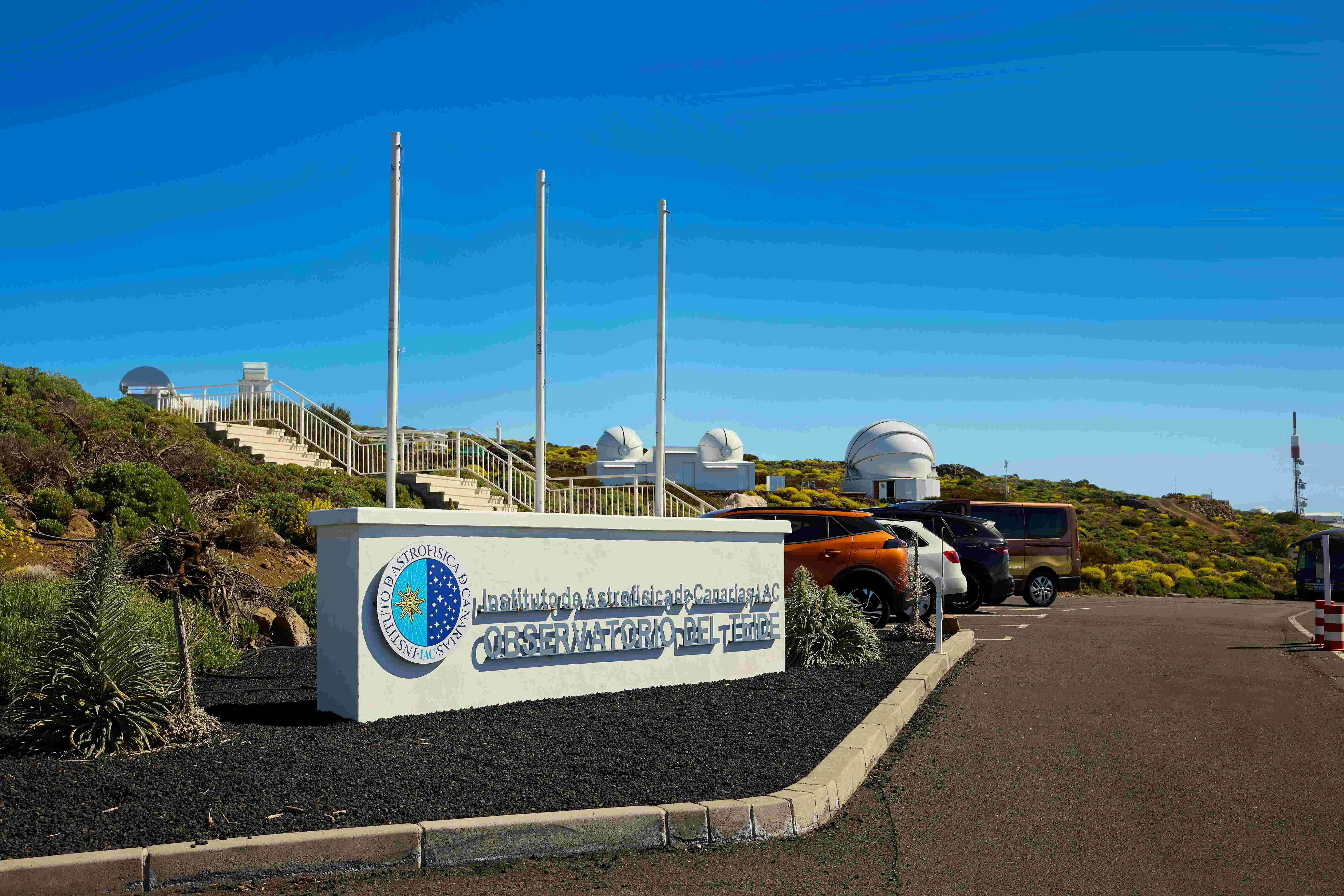 Sign reading "Observatorio del Teide" with observatory domes and parked cars in the background under a clear blue sky.
