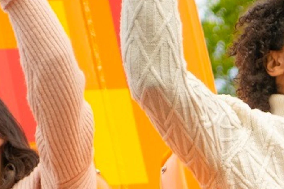 People enjoying a drop tower ride, sitting in orange seats with their arms raised, surrounded by trees and a blue sky.