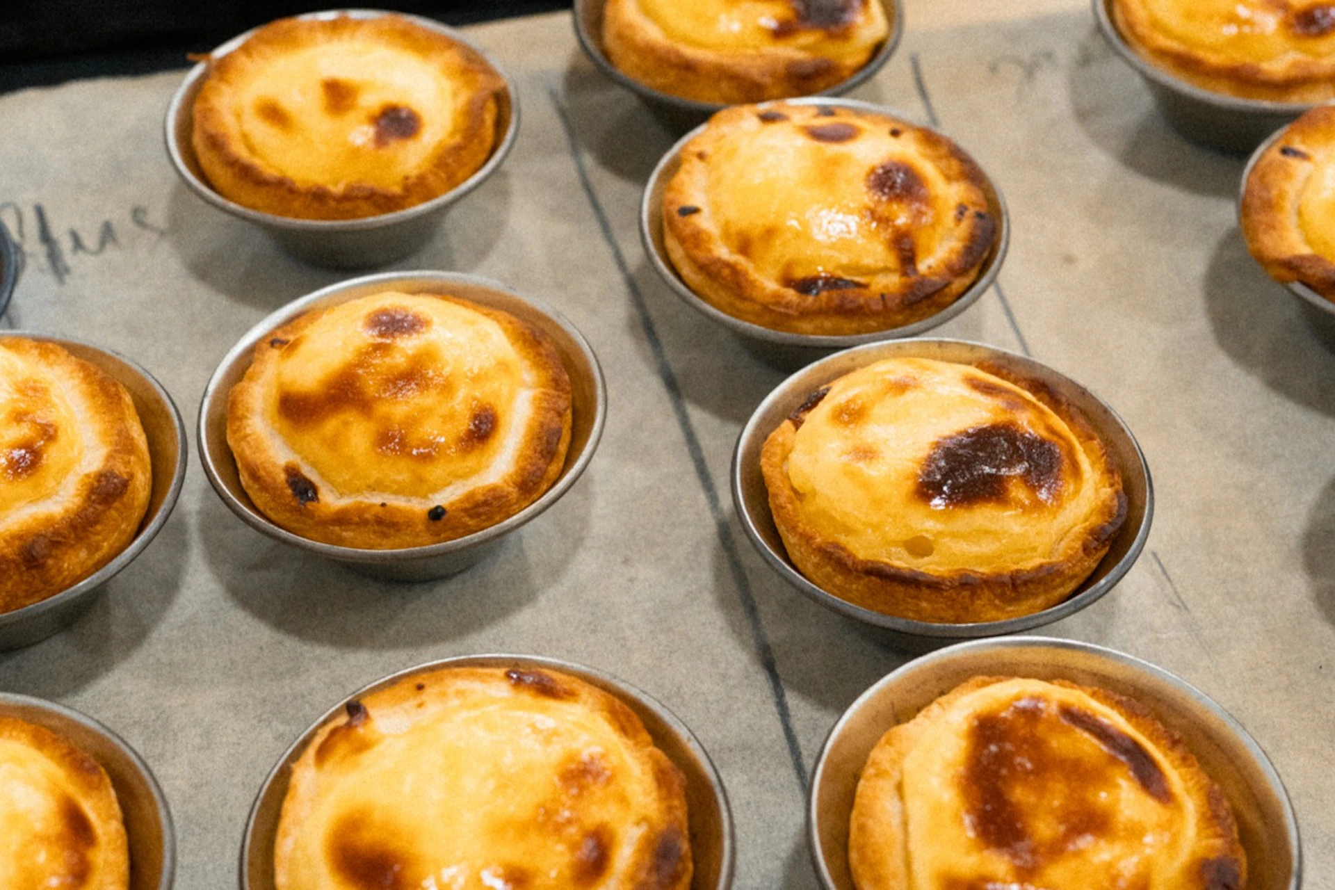 A tray of baked custard tarts in small individual tins, with golden crusts and slightly browned tops.