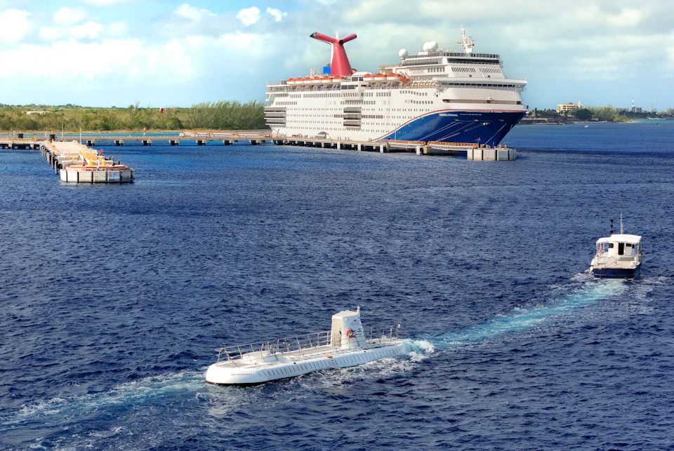 A large cruise ship is docked at a pier, with a small white submarine on the water nearby.