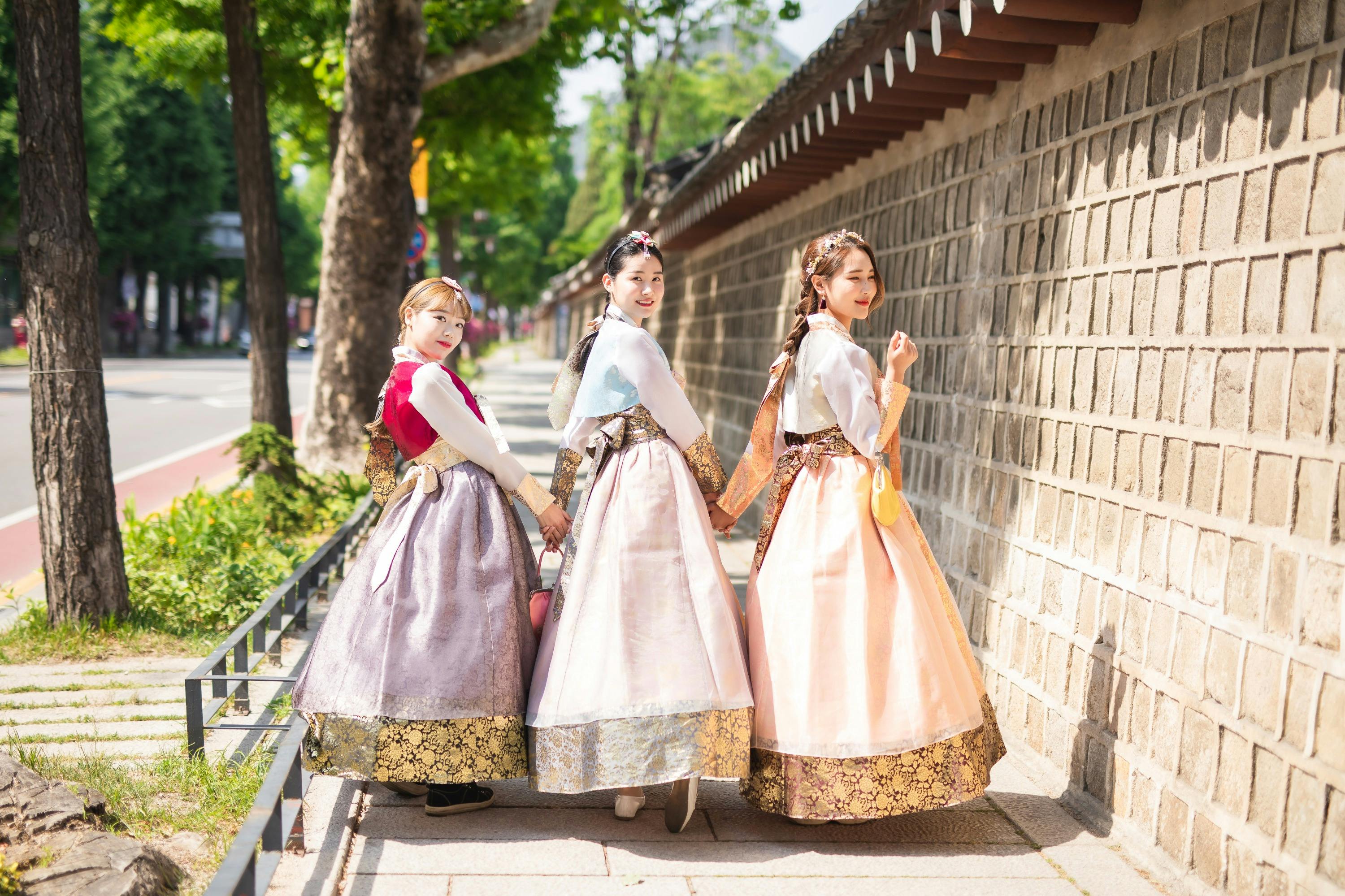 Three women in traditional Korean hanbok dresses walking along a stone wall and smiling, with greenery in the background.