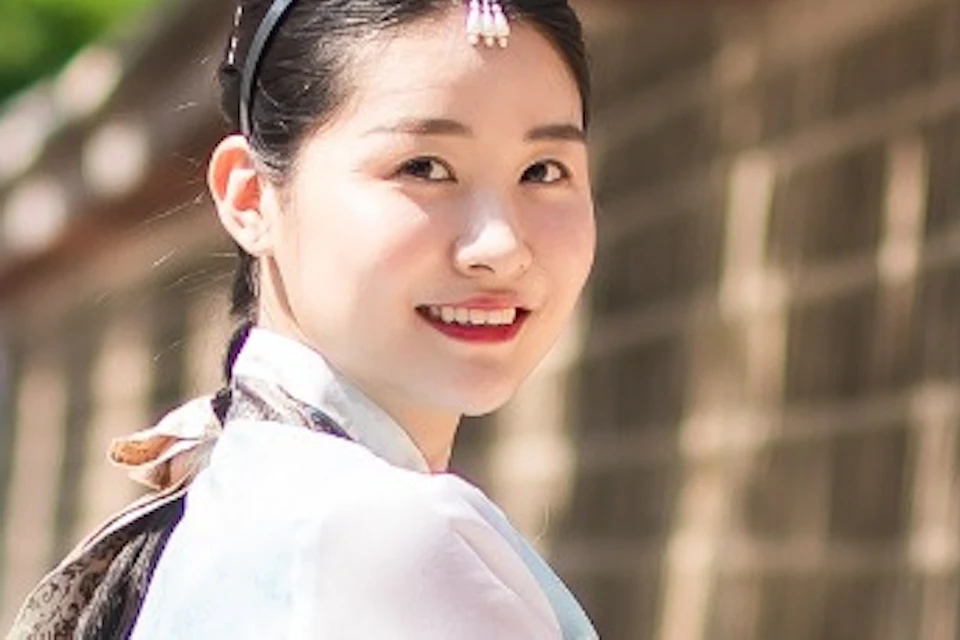 Three women in traditional Korean hanbok dresses walking along a stone wall and smiling, with greenery in the background.