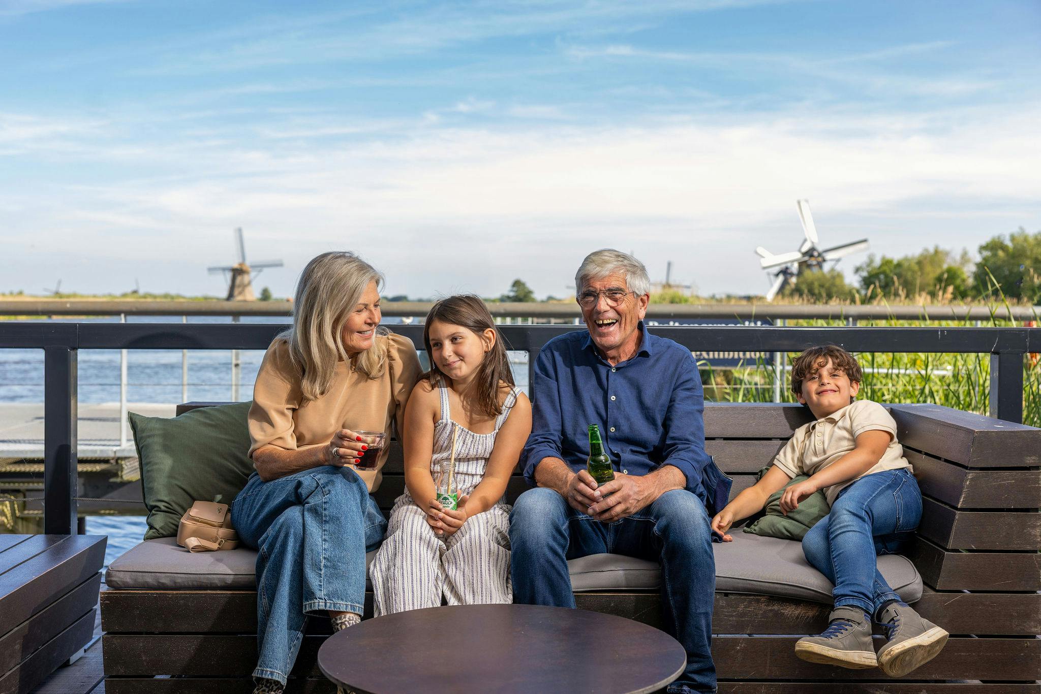 Rodina sedící venku na lavičce s větrnými mlýny v pozadí. Vypadají uvolněně a užívají si vzájemnou společnost.