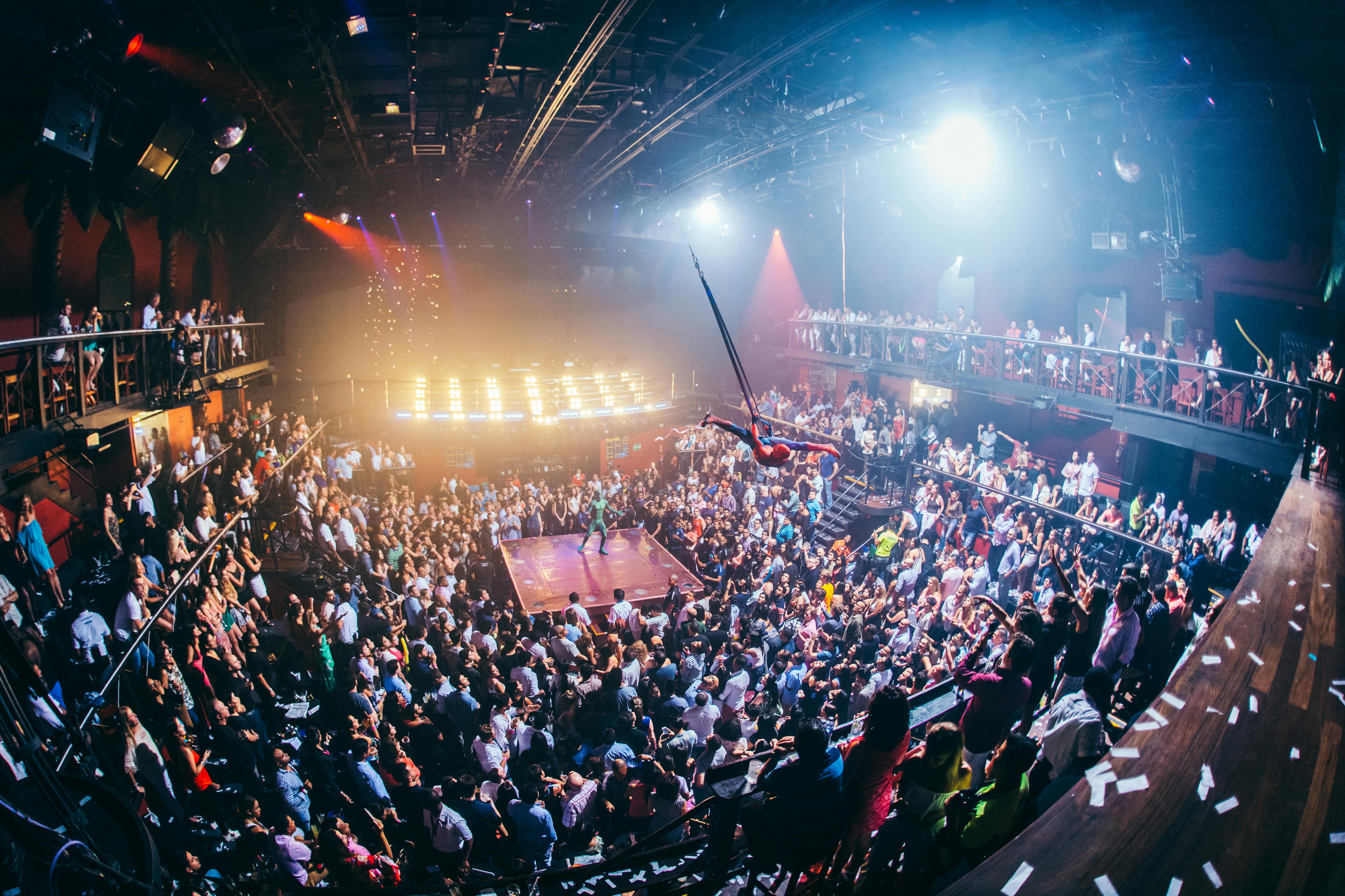 Un artista su un trapezio sopra un locale notturno affollato e su più livelli, con le luci di scena che illuminano la scena. Gli avventori guardano da diversi livelli.