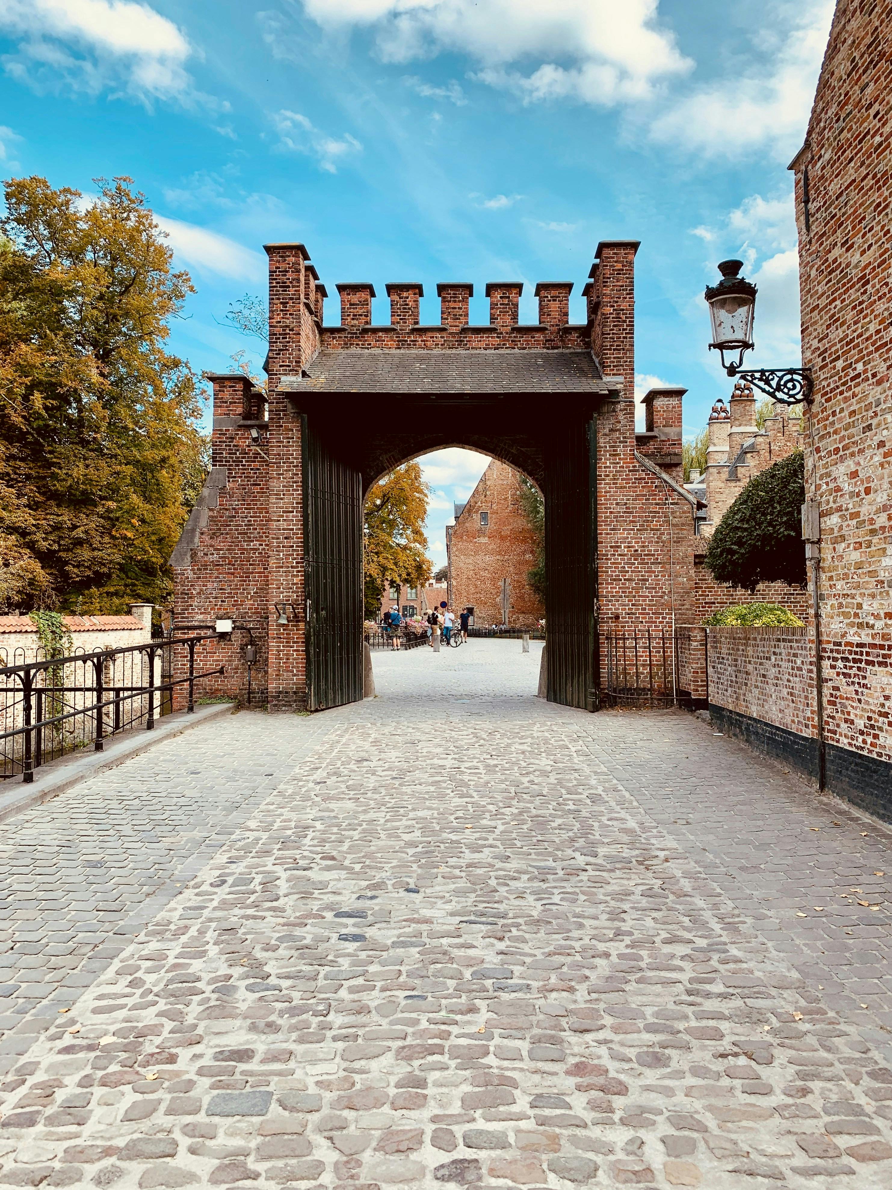 A medieval-style brick archway with crenellations over a cobblestone path, surrounded by trees and buildings.