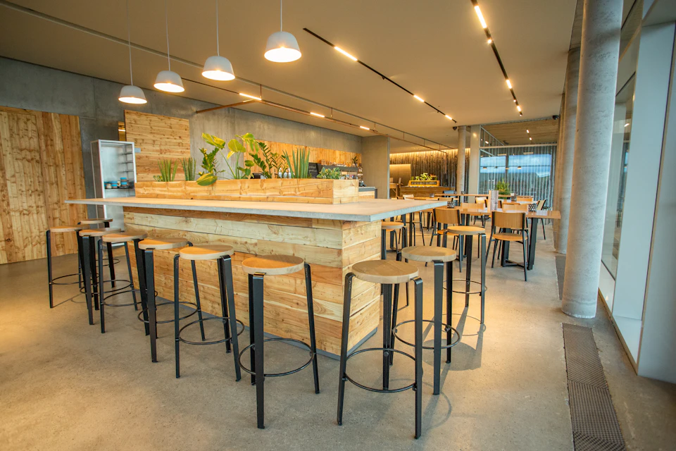 Modern cafe with wooden tables, high stools, hanging lamps, and indoor plants on a central counter. Large windows in the background.