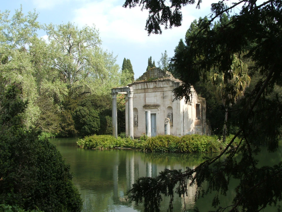 An ancient, stone temple-like structure with columns sits on an island, surrounded by a placid lake and lush trees.