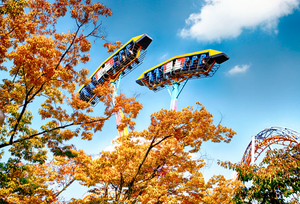 Two amusement park rides resembling large rotating platforms, surrounded by autumn trees under a blue sky.